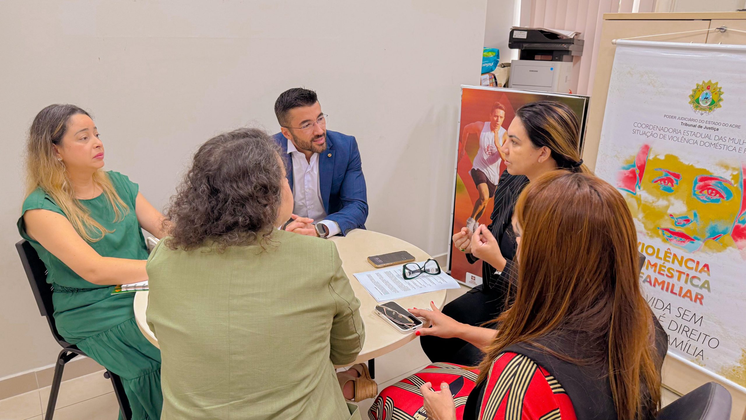 A imagem mostra um grupo de cinco pessoas, quatro mulheres e um homem, reunidas ao redor de uma mesa redonda em um ambiente interno, possivelmente uma sala de reunião ou escritório institucional. A conversa parece séria e focada, sugerindo uma discussão ou planejamento de ações, talvez relacionadas a políticas públicas ou projetos sociais. Alguns elementos reforçam essa ideia: Sobre a mesa há documentos, celulares e um par de óculos, indicando que estão em uma reunião formal. Ao fundo, há um banner parcialmente visível com a frase "Violência contra a mulher e familiar", associado à Coordenadoria da Mulher em Situação de Violência Doméstica e Familiar, o que sugere que o tema da reunião pode estar relacionado ao enfrentamento da violência doméstica. Também é visível um cartaz com a imagem de um corredor, o que pode indicar a presença de materiais de campanhas institucionais diversas no local. As pessoas estão bem vestidas e atentas, reforçando o caráter profissional e engajado da conversa.