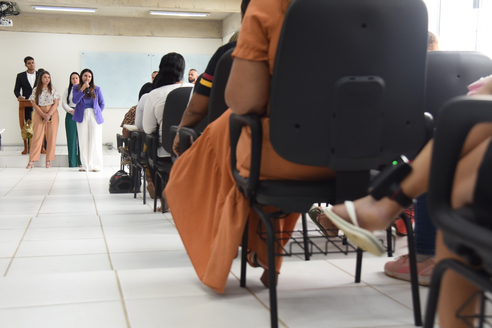 Na imagem há uma mulher com cabelos castanhos, vestindo um blazer roxo e calça branca palestrando sobre empreendedorismo em parceria com a ufac, iapen e tjac, ao lado delas estão mais duas mulheres, uma de cabelos loiros usando uma camisa branca de bolinhas e calça na cor marfim e a outra de cabelos pretos usando uma camisa branca social e calça verde. Ao fundo está um homem de terno preto e blusa social branca e em desfoco estão as mulheres assistindo a palestra.