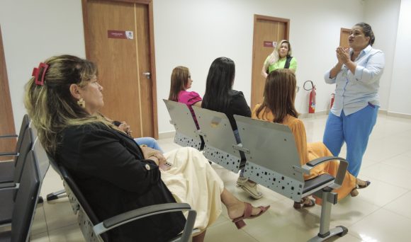 Na imagem, é possível ver a enfermeira durante a palestra, mexendo as mãos como exemplo de cuidado à saúde e prevenção. A enfermeira Ana está de frente para as mulheres, sentadas nas cadeiras metálicas, olhando para elas enquanto fala. A cena transmite a sensação de paz.