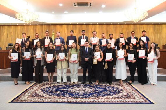 A imagem mostra um grupo de pessoas posando para uma foto oficial em um ambiente formal, provavelmente um auditório ou salão de cerimônias. O grupo é composto por homens e mulheres, organizados em duas fileiras, todos segurando certificados com capa branca e detalhes vermelhos. Ao centro da primeira fileira, está um homem de terno escuro, aparentando ser uma figura de autoridade, como um coordenador ou diretor. O fundo da imagem apresenta uma parede de madeira clara com bandeiras do Brasil ao centro, e há dois lustres elegantes no teto, sugerindo um ambiente sofisticado e institucional. O piso está coberto com um tapete azul com estampa clássica, reforçando a formalidade da ocasião. Todos estão sorrindo, indicando que se trata de um momento de celebração, como uma formatura, certificação ou encerramento de curso.