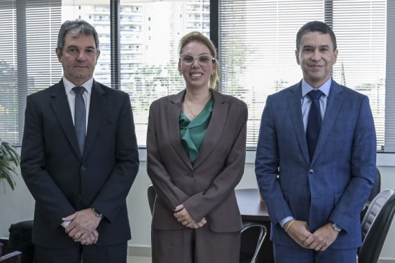 A imagem mostra três pessoas posando para uma foto em um ambiente corporativo. Os dois homens e a mulher estão vestidos formalmente, com ternos e roupas sociais. O homem à esquerda veste um terno preto com gravata cinza, enquanto o da direita usa um terno azul com gravata azul-escura. A mulher, no centro, veste um conjunto marrom com blusa verde e usa óculos. Eles estão em um escritório bem iluminado, com grandes janelas ao fundo que revelam uma paisagem urbana com prédios. As persianas estão parcialmente fechadas, e há móveis como cadeiras e uma mesa ao fundo. A expressão facial dos três é formal, mas com um leve sorriso, sugerindo um registro profissional, possivelmente de uma reunião ou evento institucional.