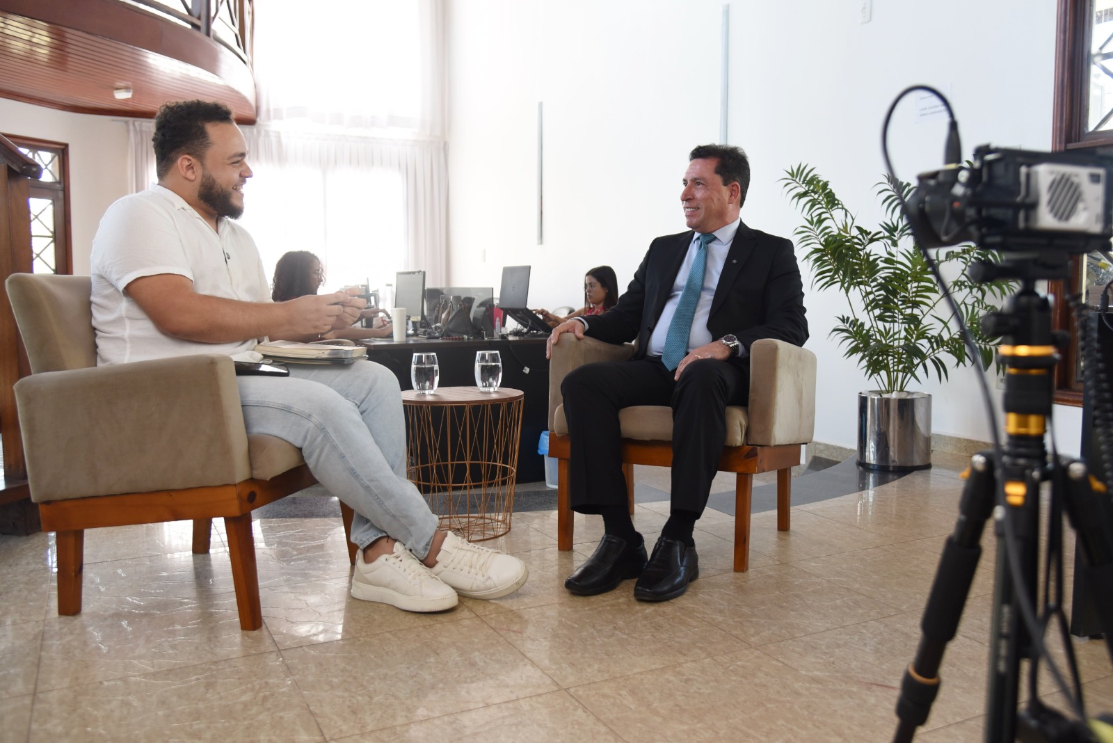 A imagem retrata uma entrevista sendo realizada em um ambiente sofisticado e bem iluminado. Dois homens estão sentados em poltronas bege de madeira, posicionadas frente a frente, com uma pequena mesa entre eles. O entrevistador, à esquerda, veste uma camisa branca casual e jeans claros, e segura um caderno e uma caneta enquanto conversa com um sorriso no rosto. Ele usa tênis brancos e parece estar conduzindo a entrevista de maneira descontraída. O entrevistado, à direita, está vestido formalmente com um terno preto, camisa branca e gravata azul. Ele mantém uma postura relaxada, com um sorriso e expressão atenta. Na mesa entre eles, há dois copos de água. Ao fundo, há um espaço de trabalho com pessoas usando laptops, além de uma planta decorativa e cortinas brancas que deixam a luz natural entrar. Na lateral direita da imagem, uma câmera profissional montada em um tripé está gravando a entrevista, indicando que se trata de uma produção audiovisual, possivelmente para um programa ou canal de comunicação.