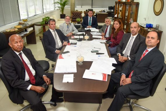 imagem mostra oito pessoas em uma sala de reunião. Todos estão sentados ao redor de uma mesa retangular, sobre a qual há alguns documentos, celulares e um microfone de mesa. Os homens ao redor da mesa vestem ternos cinza, exceto o homem à esquerda, que usa um terno preto. Eles usam gravatas nas cores cinza, azul e vermelha. O homem de terno preto e gravata vermelha, à esquerda, segura seus óculos de grau na mão. Todos estão sorrindo. A mesa contém alguns documentos, um caderno preto, uma caneta e um pequeno vaso com bombons. No fundo, há uma janela e vasos de plantas na sala.