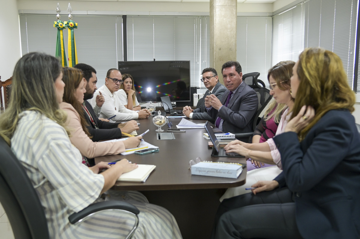 Em um ambiente de escritório bem iluminado, um grupo diversificado de profissionais do Tribunal de Justiça do Acre está reunido ao redor de uma mesa para uma reunião. Os participantes estão vestidos de forma sofisticada em trajes de negócios, e alguns seguram cadernos, enquanto outros têm laptops abertos, indicando anotações ativas ou referência a documentos importantes. A atmosfera sugere uma discussão séria, possivelmente envolvendo planejamento estratégico ou tomada de decisões, à medida que os indivíduos parecem focados e atentos às contribuições uns dos outros.