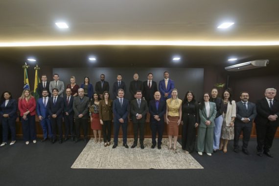 A imagem mostra um grupo de profissionais reunidos em um ambiente formal, possivelmente para um evento institucional ou solenidade oficial. O cenário inclui a presença de bandeiras do Brasil, indicando um contexto governamental ou jurídico.