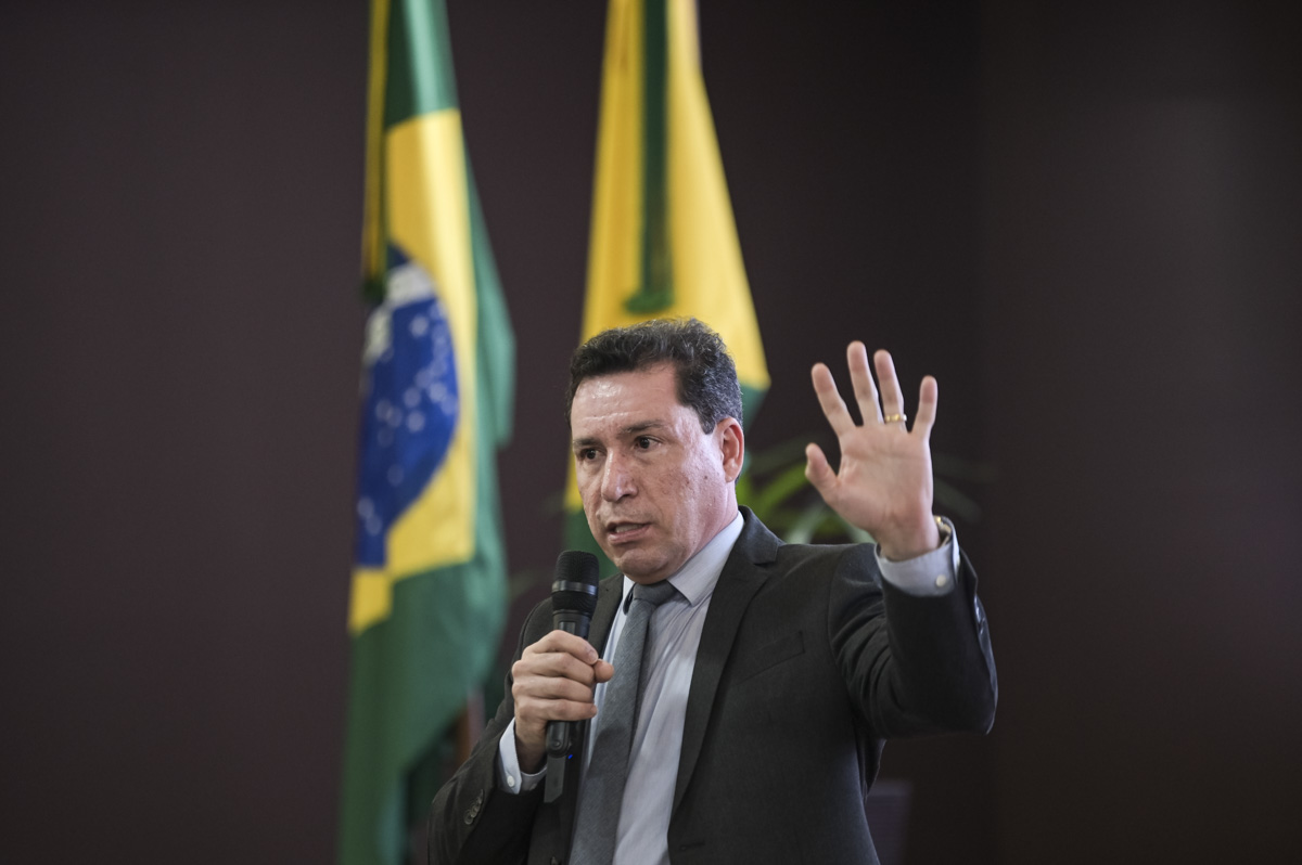 A imagem mostra um homem de terno segurando um microfone e discursando em um evento oficial. Ao fundo, há bandeiras do Brasil e possivelmente de um estado brasileiro, sugerindo que a ocasião é institucional. Pelo contexto das imagens anteriores, é provável que ele esteja participando de um evento do Poder Judiciário do Acre