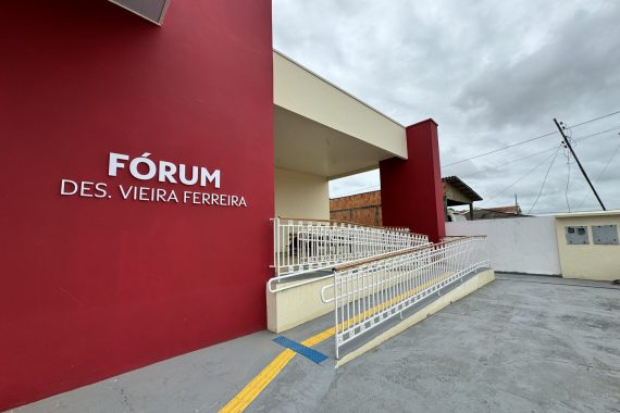 A imagem mostra a fachada de um edifício institucional identificado como Fórum Des. Vieira Ferreira. A construção possui uma parede externa em tom vermelho escuro, com o nome do fórum escrito em letras brancas em destaque. À frente do prédio, há uma rampa de acesso com corrimãos brancos, sinalização tátil e piso antiderrapante, indicando acessibilidade para pessoas com mobilidade reduzida. A estrutura é moderna, com um design simples e funcional. O céu está nublado, com nuvens densas, sugerindo que o clima está encoberto ou próximo de chuva. Ao fundo, é possível ver parte de edificações residenciais e fiações elétricas, indicando que o fórum está localizado em uma área urbana.