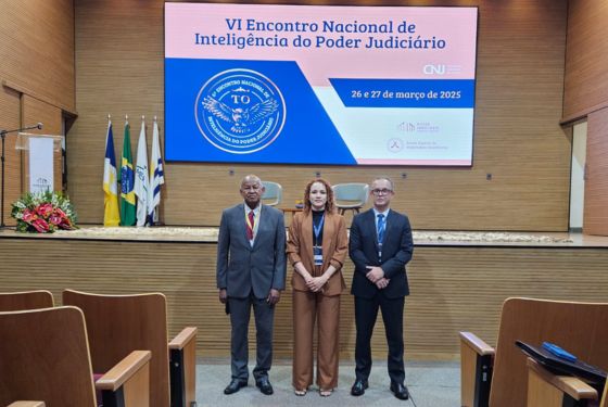 A imagem mostra seis pessoas reunidas em um auditório grande atras do mesmo o palco com um par de cadeira e um telão grande com informações na tela sobre o encontro nacional de inteligência artificial Todos participarão e ambos possando para a foto sorrindo, os homens de terno a combina apenas dois de gravatas vermelha, e o outro azul já a mulher de blusa preto com blazer marrom e calça marrom sapato preto. s pessoas na foto estão sorrindo a iluminação é clara, vinda as luzes do teto.