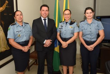 A imagem mostra quatro pessoas posando para uma foto em um ambiente institucional, provavelmente relacionado à segurança pública ou à administração pública. Ao centro está um homem de terno escuro, camisa branca e gravata azul, sorrindo para a câmera. Ele está ladeado por três mulheres fardadas da Polícia Militar, com uniformes azuis e insígnias que indicam patentes superiores. Duas delas usam óculos e todas estão igualmente sorrindo, demonstrando um momento de cordialidade e formalidade. Ao fundo, há duas bandeiras do Brasil e do estado (possivelmente do Amapá, considerando o contexto anterior), além de uma pintura clássica da deusa da justiça com uma balança, reforçando o ambiente institucional e jurídico. Também é possível ver parte de uma televisão ou tela de apresentação à direita. A imagem transmite um momento oficial, possivelmente uma reunião ou reconhecimento institucional entre autoridades civis e militares.