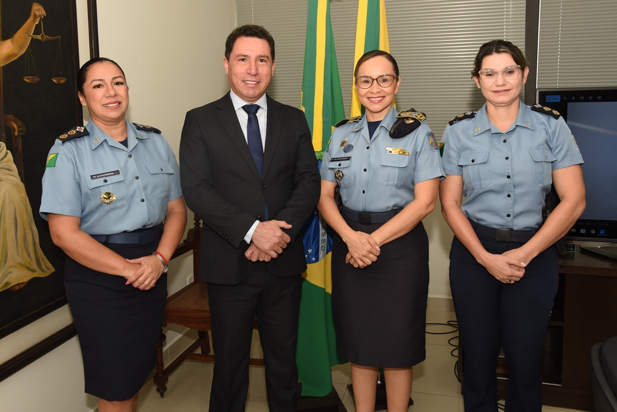 A imagem mostra quatro pessoas posando para uma foto em um ambiente institucional, provavelmente relacionado à segurança pública ou à administração pública. Ao centro está um homem de terno escuro, camisa branca e gravata azul, sorrindo para a câmera. Ele está ladeado por três mulheres fardadas da Polícia Militar, com uniformes azuis e insígnias que indicam patentes superiores. Duas delas usam óculos e todas estão igualmente sorrindo, demonstrando um momento de cordialidade e formalidade. Ao fundo, há duas bandeiras do Brasil e do estado (possivelmente do Amapá, considerando o contexto anterior), além de uma pintura clássica da deusa da justiça com uma balança, reforçando o ambiente institucional e jurídico. Também é possível ver parte de uma televisão ou tela de apresentação à direita. A imagem transmite um momento oficial, possivelmente uma reunião ou reconhecimento institucional entre autoridades civis e militares.