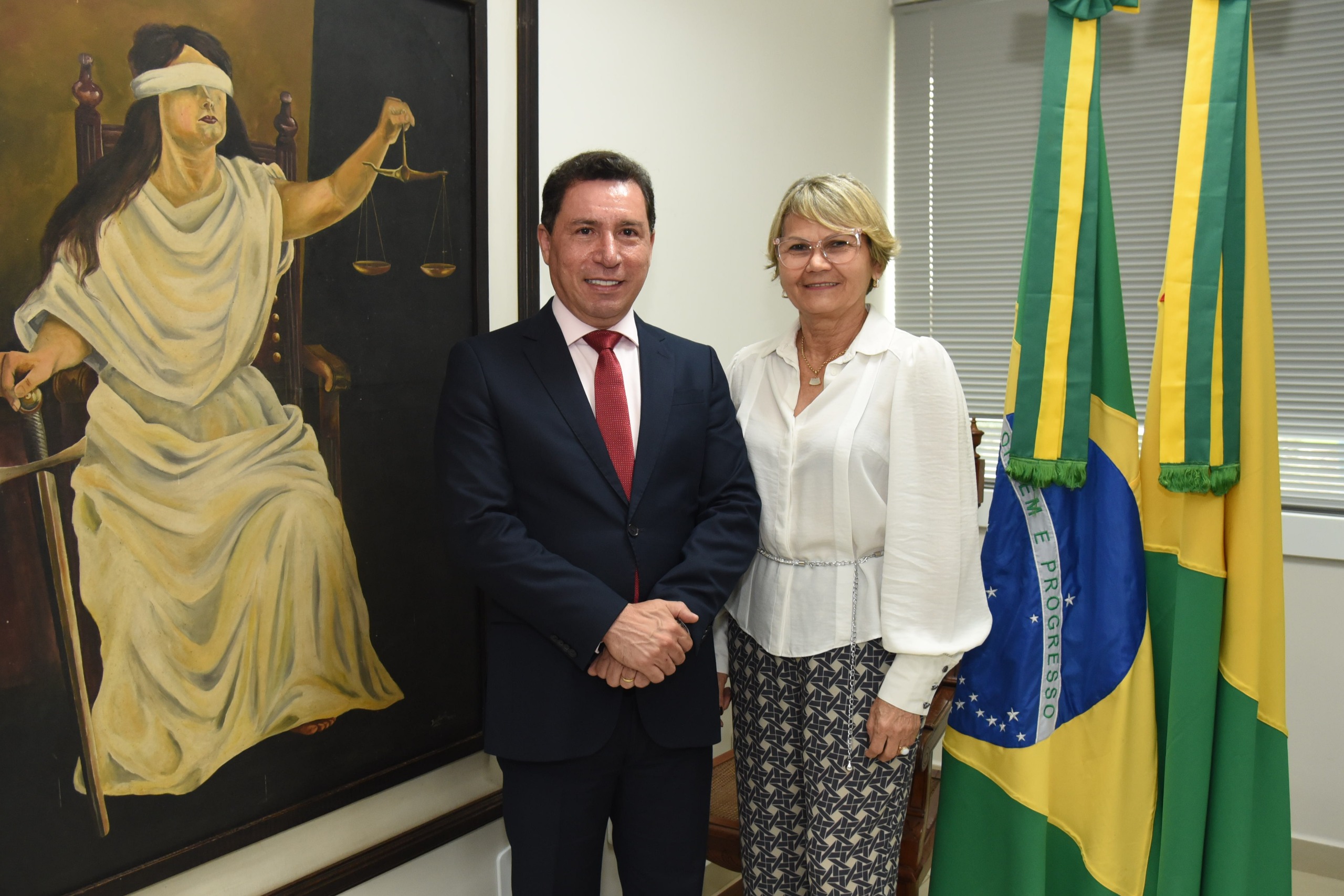 A imagem mostra um homem e uma mulher posando lado a lado para uma foto em um ambiente formal. O homem está vestindo um terno escuro com gravata vermelha, enquanto a mulher usa uma blusa branca e uma saia ou calça estampada. Ao fundo, há uma grande pintura da figura da Justiça, representada como uma mulher vendada segurando uma balança e uma espada, um símbolo tradicional do sistema judiciário. Além disso, há duas bandeiras do Brasil posicionadas ao lado da dupla. O ambiente sugere um escritório ou gabinete institucional, possivelmente relacionado ao poder judiciário ou a alguma repartição pública. Ambos os indivíduos aparentam estar em um evento ou encontro formal, possivelmente relacionado ao contexto jurídico ou governamental.