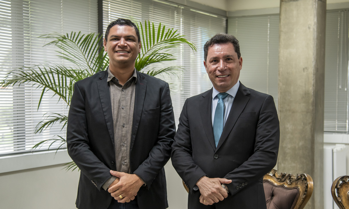 A imagem mostra dois homens posando para uma foto em um ambiente corporativo. Ambos estão bem vestidos e sorriem para a câmera. O homem à esquerda usa um blazer preto sobre uma camisa social cinza escuro, enquanto o homem à direita veste um terno preto com gravata azul. O fundo da imagem inclui uma planta decorativa, janelas com persianas e um pilar de concreto, sugerindo um escritório ou sala institucional. Também é possível ver parte de uma poltrona de madeira estofada, reforçando o ambiente formal.