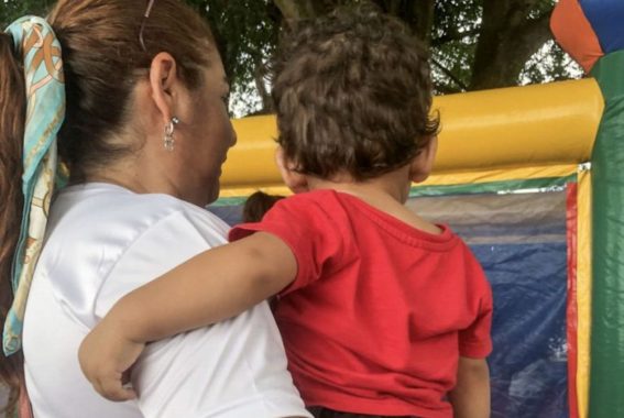 A imagem mostra uma mulher carregando uma criança no colo. A mulher está de costas, usando uma blusa branca e tem os cabelos presos com um lenço azul com detalhes claros. Ela também usa brincos e óculos de leitura apoiados na cabeça. A criança, que também está de costas, veste uma camiseta vermelha e tem cabelos castanhos encaracolados. Ao fundo, há uma estrutura colorida inflável, provavelmente um brinquedo como um pula-pula ou castelinho, indicando que eles podem estar em um evento ao ar livre, como uma festa ou feira infantil. Há árvores ao fundo, sugerindo que o local é arborizado.