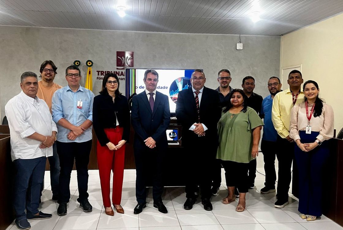 A imagem mostra um grupo de 14 pessoas posando para uma foto em ambiente institucional, possivelmente após uma reunião ou evento oficial. O local é identificado como parte do Tribunal de Justiça, com a palavra "TRIBUNAL" e o brasão do Poder Judiciário visíveis ao fundo, junto a uma bandeira. As pessoas estão organizadas em duas fileiras: uma mais à frente, composta por indivíduos em trajes formais (terno, blazers, roupas sociais), e outra ao fundo, com roupas um pouco mais casuais, embora ainda adequadas ao ambiente de trabalho. Ao centro da imagem, há uma tela exibindo uma apresentação com o título "Gestão de Produtividade", sugerindo que o encontro está relacionado à avaliação de desempenho ou planejamento institucional. O grupo parece composto por servidores públicos, gestores e colaboradores de diferentes áreas, evidenciando um momento de integração, capacitação ou celebração de resultados.