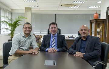 Na imagem, vemos um homem vestido com uma camisa verde clara e calça de tom pastel claro; outro homem está com paletó preto, calça preta, blusa por dentro do paletó preto e gravata azul listrada com detalhes em cinza e branco; já o outro homem, ao lado, está com blazer preto, blusa azul escura e calça preta. Os três homens estão com as mãos sobre a mesa, se apoiando e sorrindo para a câmera. A imagem transmite a sensação de alegria e harmonia no ambiente.
