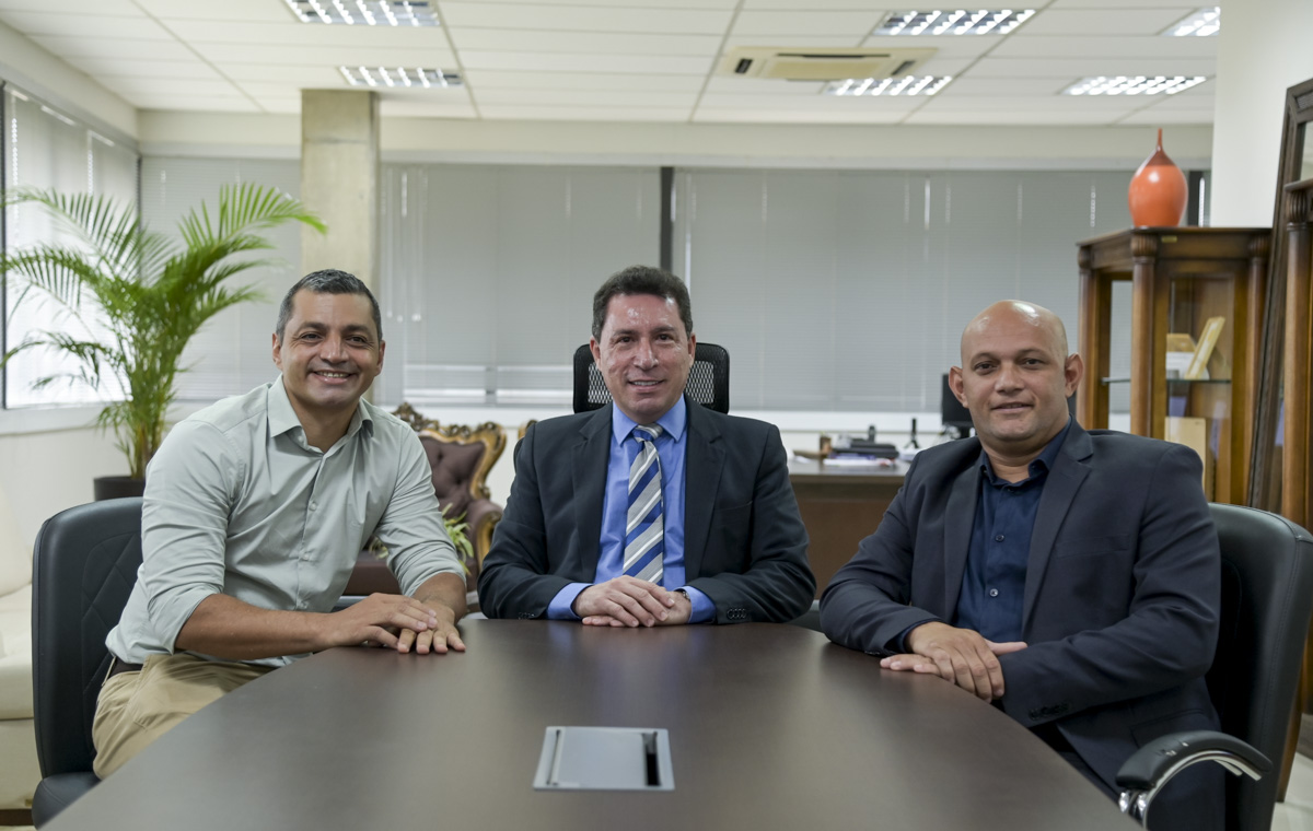 Na imagem, vemos um homem vestido com uma camisa verde clara e calça de tom pastel claro; outro homem está com paletó preto, calça preta, blusa por dentro do paletó preto e gravata azul listrada com detalhes em cinza e branco; já o outro homem, ao lado, está com blazer preto, blusa azul escura e calça preta. Os três homens estão com as mãos sobre a mesa, se apoiando e sorrindo para a câmera. A imagem transmite a sensação de alegria e harmonia no ambiente.