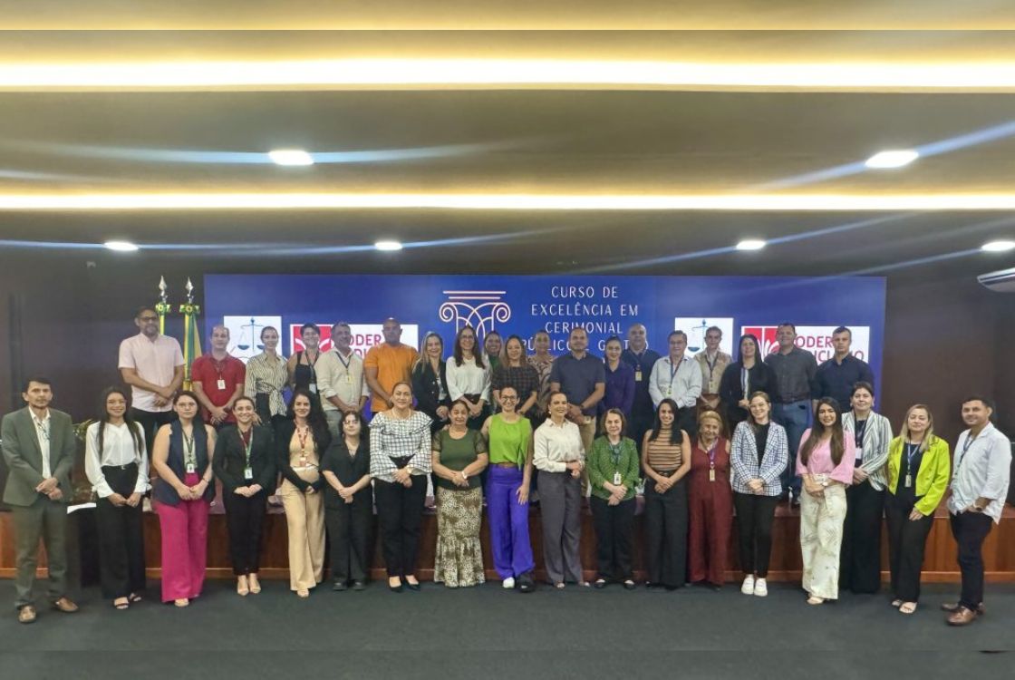 A imagem mostra um grupo de cerca de 40 pessoas posando para uma foto em um auditório, durante o “Curso de Excelência em Cerimonial Público e Gestão”. Elas estão vestidas de maneira profissional e variada, com roupas formais e semi formal, divididas em duas fileiras, à frente e sobre um palco. O fundo tem um painel azul com o nome do curso e logotipos do Poder Judiciário. Todos estão vestidos de forma profissional e demonstram expressões amigáveis.