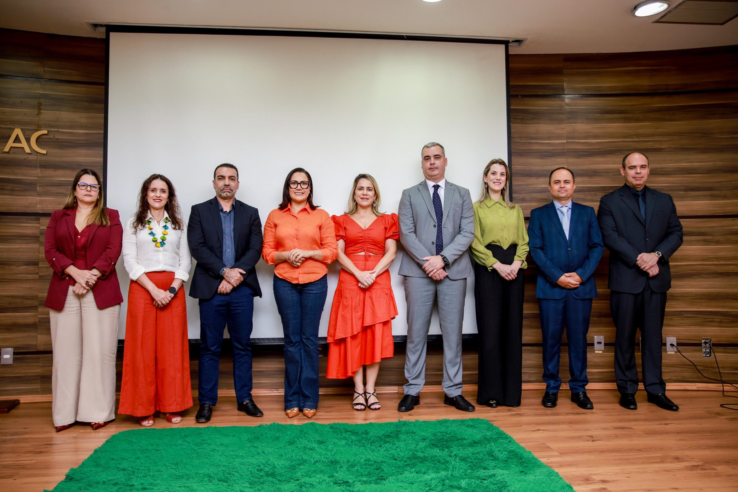 A imagem mostra nove pessoas posicionadas lado a lado, de forma organizada, em frente a um telão branco em uma sala com painéis de madeira nas paredes. O grupo está formalmente vestido, sugerindo a participação em um evento institucional ou cerimônia oficial. As mulheres usam roupas sociais, algumas com blusas coloridas e saias ou calças elegantes, enquanto os homens estão de terno e gravata. A disposição, o fundo neutro e o tapete verde à frente do grupo reforçam o tom solene da ocasião. Ao fundo, à esquerda da imagem, é possível ver parte das letras "AC", o que indica que o evento pode estar relacionado ao estado do Acre. A expressão corporal dos participantes transmite formalidade e respeito, com todos olhando para a câmera em pose oficial.