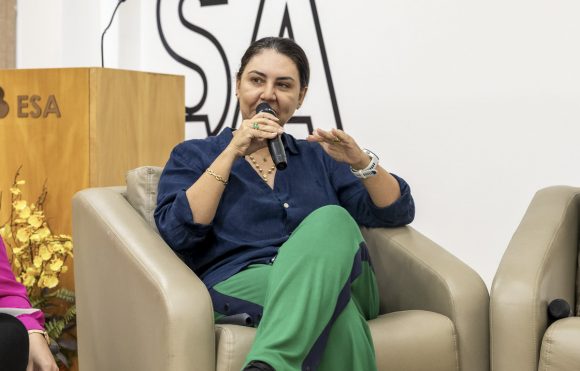 A imagem mostra uma mulher sentada em uma poltrona bege, falando ao microfone em um ambiente interno que parece ser um auditório ou sala de eventos. Ao fundo, vemos novamente a sigla "ESA" na parede, reforçando que o local está relacionado à Escola Superior da Advocacia ou a alguma instituição com essas iniciais. A mulher veste uma camisa azul escura e uma calça verde com detalhes em azul escuro nas laterais. Ela usa acessórios como pulseiras douradas, um colar, brincos pequenos, um anel verde e um relógio branco no pulso esquerdo. Seu cabelo está preso para trás, e ela parece estar engajada na fala, gesticulando com a mão esquerda enquanto segura o microfone com a direita. À esquerda da imagem, parcialmente visível, está o braço de outra pessoa, sugerindo que essa fala pode fazer parte de um painel ou roda de conversa. Há também um arranjo floral amarelo decorando o canto do ambiente e um púlpito de madeira com a sigla "ESA" gravada.