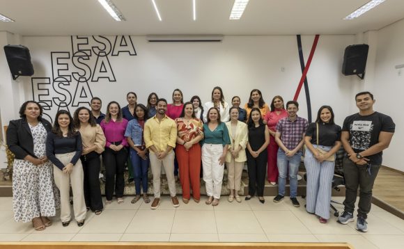A imagem mostra um grupo de aproximadamente 26 pessoas posando para uma foto em um ambiente fechado, provavelmente uma sala de eventos ou auditório. Há uma parede branca ao fundo com a sigla "ESA" repetida várias vezes em letras grandes e pretas, o que pode indicar que o local pertence à Escola Superior da Advocacia (ou instituição com sigla semelhante). As pessoas estão organizadas em duas fileiras: uma fileira em pé na frente e outra atrás, ligeiramente elevada em uma plataforma. A maioria está sorrindo e usando roupas casuais ou sociais, com uma variedade de estilos e cores — algumas vestindo roupas mais formais como blazers e calças de alfaiataria, enquanto outras optam por roupas mais descontraídas. O ambiente é bem iluminado com luzes fluorescentes e há alto-falantes montados nas paredes. No lado direito da imagem, veem-se duas faixas verticais nas cores preta e vermelha que descem da parede para o chão, possivelmente parte da decoração institucional. Parece ser uma foto tirada ao final de um evento, palestra ou curso, celebrando um momento coletivo do grupo.