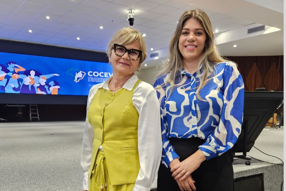 A imagem mostra duas mulheres em pé, lado a lado, posando para uma foto em um ambiente interno que parece ser um auditório moderno, possivelmente durante um evento institucional. À esquerda, está uma mulher de cabelos curtos e loiros, usando óculos grandes e escuros. Ela veste uma camisa branca por baixo de um colete e saia verde-limão, transmitindo um estilo elegante e profissional. À direita, está uma mulher de cabelos longos e loiros, soltos. Ela veste uma camisa azul com estampa branca abstrata e uma saia ou calça preta. Também sorri levemente, transmitindo simpatia e confiança. Ao fundo, há um telão azul com a palavra "COCEV" e o logotipo da instituição, além de ilustrações de pessoas apontando para frente, sugerindo uma mensagem de liderança, futuro ou protagonismo. A ambientação, com teto de forro rebaixado e iluminação embutida, reforça o caráter corporativo ou acadêmico do local.