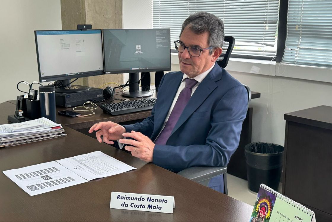 A imagem mostra um homem em um ambiente de escritório, identificado por uma placa com o nome "Raimundo Nonato da Costa Maia", vestindo terno azul, camisa branca e gravata roxa. Ele está sentado à mesa, gesticulando com as mãos, sugerindo que está em uma reunião ou explicando algo durante uma conversa. Sobre a mesa, há documentos, um calendário de mesa, e itens organizacionais como porta-canetas e uma garrafa térmica. Dois monitores de computador estão à sua frente, e um dos desktops exibe a logomarca do Poder Judiciário, o que indica que se trata de um ambiente institucional, possivelmente ligado ao setor jurídico ou administrativo de um tribunal. O ambiente é bem iluminado, com janelas com persianas parcialmente fechadas ao fundo. A postura do homem e a formalidade do local reforçam o contexto profissional, possivelmente uma entrevista, apresentação de resultados ou momento de planejamento estratégico.
