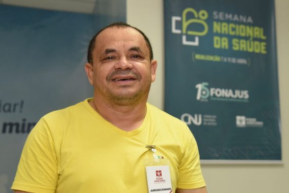 A imagem mostra um momento em que o homem esta possando pra foto sorrindo, o mesmo tem crachá de identificação e ao lado dele um banner com a seguinte informação: Semana nacional da saúde. O ambiente transmite o foco no bem-estar e saúde.