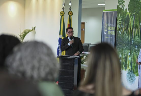 A imagem mostra um homem de terno escuro e gravata azul clara falando ao microfone em um ambiente formal. Ele está posicionado atrás de um púlpito, onde repousam alguns papéis ou documentos. O homem parece estar fazendo um discurso ou pronunciamento oficial. Ao fundo, há duas bandeiras nas cores verde, amarelo e vermelho, identificando que o evento provavelmente acontece no estado do Acre, Brasil. Também é possível ver plantas decorativas ao redor, sugerindo um ambiente bem organizado, possivelmente dentro de uma instituição pública ou governamental. O foco está no homem discursando, enquanto outras pessoas aparecem desfocadas no primeiro plano, indicando a presença de uma plateia. A cena transmite uma atmosfera institucional e solene.
