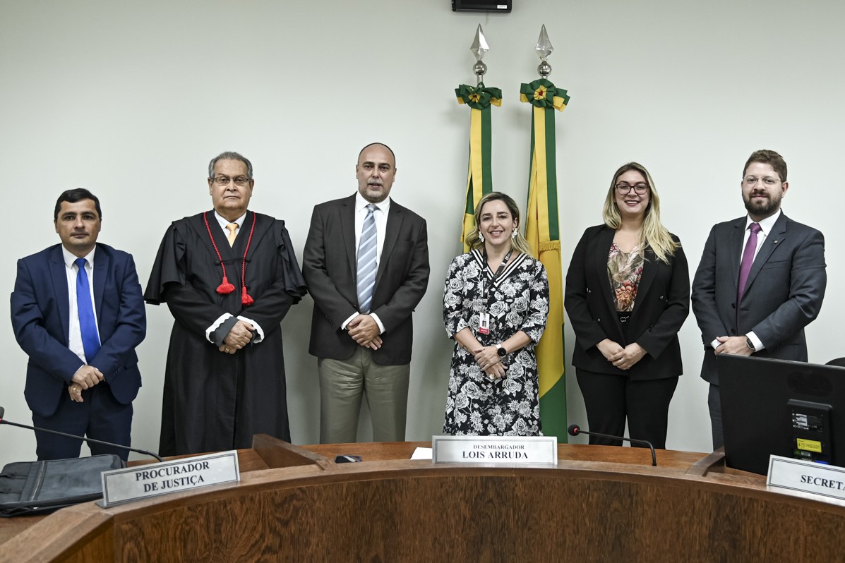 A imagem mostra um grupo de seis pessoas em pé, alinhadas lado a lado, posando para uma foto formal em um ambiente institucional. Ao fundo, há duas bandeiras com as cores verde e amarela, típicas do Brasil, indicando que o evento pode estar relacionado ao setor público ou judiciário. As pessoas estão vestidas de forma formal, com ternos e vestidos sociais. Um dos homens, o segundo da esquerda para a direita, está usando uma toga com detalhes em vermelho, sinalizando que provavelmente é um magistrado. Diante das pessoas, há uma bancada de madeira em formato curvo, com placas de identificação. A placa central exibe o nome "Lois Arruda", com o título de "Desembargador", sugerindo que ele é um dos participantes principais da sessão. À esquerda, vê-se uma placa com o título "Procurador de Justiça", enquanto à direita aparece outra com a palavra "Secret...". A cena aparenta ser de uma sessão ou reunião oficial, possivelmente em um tribunal ou órgão do Ministério Público. Se quiser, posso ajudar a criar uma legenda formal ou um release institucional a partir dessa imagem.