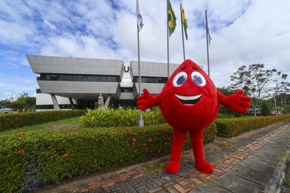 A imagem mostra uma figura fantasiosa em formato de gota de sangue sorridente, com grandes olhos azuis e braços abertos, posando alegremente em frente a um prédio moderno de arquitetura arrojada. O personagem provavelmente representa uma campanha de doação de sangue ou promoção da saúde, transmitindo uma mensagem de otimismo e incentivo à participação da população. Ao lado direito da imagem, há uma estátua de uma mulher de pé, trajando vestes formais e segurando livros, representando simbolicamente a Justiça, a sabedoria ou o conhecimento. Na base da estátua, há uma placa com inscrições, parcialmente visível. O cenário é bem cuidado, com canteiros floridos e arbustos aparados, e o ambiente transmite uma sensação de organização institucional. O prédio ao fundo, com sua fachada espelhada e linhas geométricas, sugere se tratar de uma instituição pública ou governamental.