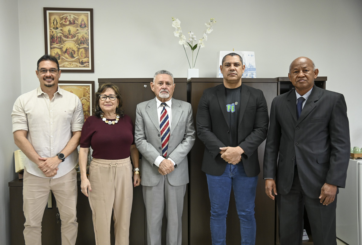 A imagem mostra cinco pessoas posando para uma foto em um ambiente interno, provavelmente um escritório ou repartição pública. O grupo é composto por três homens e uma mulher, todos posicionados lado a lado, de frente para a câmera. Da esquerda para a direita: Um homem com camisa clara de manga longa e calça bege, sorrindo. Uma mulher com blusa vinho, colar branco e calça clara, também sorrindo. Um homem mais velho com cabelo grisalho, vestindo terno cinza claro e gravata listrada, com expressão séria. Um homem de camiseta preta com estampa "FE", blazer preto e calça jeans azul, com expressão neutra. Um homem de terno escuro e gravata azul, com expressão séria. Ao fundo, há armários de madeira escura, sobre os quais estão objetos decorativos como um vaso com flores brancas, papéis e uma pequena escultura. Na parede, há dois quadros religiosos e, à direita, uma pequena mesa com garrafas térmicas, copos e uma geladeira branca. A atmosfera da imagem é formal, sugerindo que se trata de um encontro institucional, possivelmente ligado à administração pública, educação ou sistema judiciário.