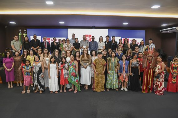 A imagem retrata um grande grupo de pessoas reunidas para uma foto oficial em um auditório institucional. O pano de fundo exibe os logotipos do Poder Judiciário do Estado do Amazonas e da ESA (Escola Superior da Advocacia), indicando um evento formal com foco educacional, jurídico ou cultural. A diversidade do grupo é um destaque marcante da foto: estão presentes representantes de comunidades indígenas — identificáveis por seus trajes tradicionais, cocares, colares e estampas culturais — juntamente com outras pessoas vestidas de forma formal ou casual, o que evidencia um encontro inclusivo e plural. Entre os participantes, há mulheres, homens, crianças e adolescentes, todos reunidos de maneira organizada, distribuídos em várias fileiras para a foto. A atmosfera transmite respeito, união e celebração da diversidade cultural e étnica, possivelmente em um contexto de valorização dos povos originários ou de direitos humanos.