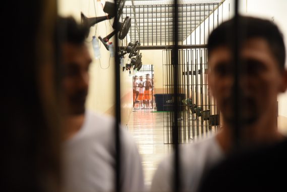 Uma imagem mostra um corredor de prisão com grades, onde vários detentos estão presos. A frente se encontra duas pessoas com camisa brancas em desfoco.