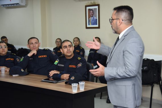 A imagem mostra uma palestra ou treinamento sendo ministrado em uma sala de situação e gerenciamento, voltada possivelmente a agentes de segurança pública. Descrição da cena: Um homem de terno cinza claro, óculos e gravata está em pé, gesticulando enquanto fala para um grupo de pessoas uniformizadas, indicando que é o palestrante ou facilitador da atividade. Os participantes estão sentados ao redor de uma mesa longa e retangular, usando uniformes escuros com insígnias e bandeiras do Brasil nos braços, sugerindo que são membros de alguma corporação — provavelmente policiais militares ou bombeiros. Sobre a mesa há copos descartáveis, garrafas de água e bonés do uniforme, indicando que é uma sessão relativamente longa e informal. Ao fundo, uma grande tela exibe uma apresentação com os termos “Mundo VUCA” e “Mundo BANI”, conceitos usados para descrever contextos modernos de instabilidade, incerteza e complexidade, bastante aplicados em ambientes de liderança e estratégia. Parte da frase visível na tela diz: “Como podemos definir ou entender nossa realidade atual?”, o que sugere que o tema da palestra está relacionado a cenários contemporâneos, tomada de decisão e adaptação em tempos de mudança. Ambiente: A sala possui acabamento moderno, com painéis de madeira e ar-condicionado. O letreiro ao fundo indica que é a “Sala de Situação e Gerenciamento”, reforçando o caráter tático e estratégico da reunião.