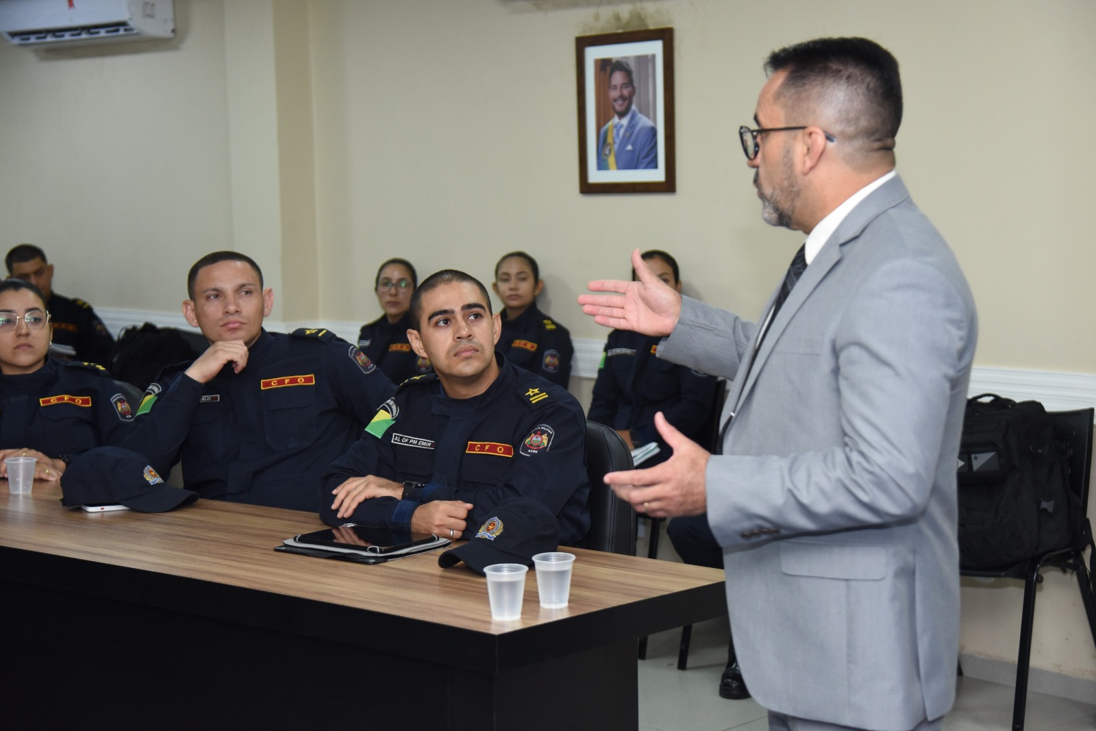 A imagem mostra uma palestra ou treinamento sendo ministrado em uma sala de situação e gerenciamento, voltada possivelmente a agentes de segurança pública. Descrição da cena: Um homem de terno cinza claro, óculos e gravata está em pé, gesticulando enquanto fala para um grupo de pessoas uniformizadas, indicando que é o palestrante ou facilitador da atividade. Os participantes estão sentados ao redor de uma mesa longa e retangular, usando uniformes escuros com insígnias e bandeiras do Brasil nos braços, sugerindo que são membros de alguma corporação — provavelmente policiais militares ou bombeiros. Sobre a mesa há copos descartáveis, garrafas de água e bonés do uniforme, indicando que é uma sessão relativamente longa e informal. Ao fundo, uma grande tela exibe uma apresentação com os termos “Mundo VUCA” e “Mundo BANI”, conceitos usados para descrever contextos modernos de instabilidade, incerteza e complexidade, bastante aplicados em ambientes de liderança e estratégia. Parte da frase visível na tela diz: “Como podemos definir ou entender nossa realidade atual?”, o que sugere que o tema da palestra está relacionado a cenários contemporâneos, tomada de decisão e adaptação em tempos de mudança. Ambiente: A sala possui acabamento moderno, com painéis de madeira e ar-condicionado. O letreiro ao fundo indica que é a “Sala de Situação e Gerenciamento”, reforçando o caráter tático e estratégico da reunião.