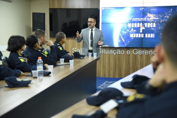 A imagem mostra uma palestra ou treinamento sendo ministrado em uma sala de situação e gerenciamento, voltada possivelmente a agentes de segurança pública. Descrição da cena: Um homem de terno cinza claro, óculos e gravata está em pé, gesticulando enquanto fala para um grupo de pessoas uniformizadas, indicando que é o palestrante ou facilitador da atividade. Os participantes estão sentados ao redor de uma mesa longa e retangular, usando uniformes escuros com insígnias e bandeiras do Brasil nos braços, sugerindo que são membros de alguma corporação — provavelmente policiais militares ou bombeiros. Sobre a mesa há copos descartáveis, garrafas de água e bonés do uniforme, indicando que é uma sessão relativamente longa e informal. Ao fundo, uma grande tela exibe uma apresentação com os termos “Mundo VUCA” e “Mundo BANI”, conceitos usados para descrever contextos modernos de instabilidade, incerteza e complexidade, bastante aplicados em ambientes de liderança e estratégia. Parte da frase visível na tela diz: “Como podemos definir ou entender nossa realidade atual?”, o que sugere que o tema da palestra está relacionado a cenários contemporâneos, tomada de decisão e adaptação em tempos de mudança. Ambiente: A sala possui acabamento moderno, com painéis de madeira e ar-condicionado. O letreiro ao fundo indica que é a “Sala de Situação e Gerenciamento”, reforçando o caráter tático e estratégico da reunião.
