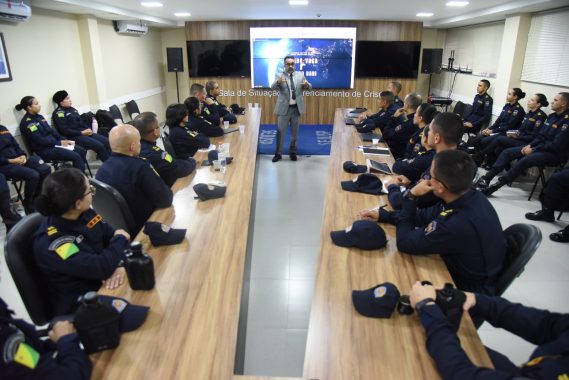 A imagem mostra uma palestra ou treinamento sendo ministrado em uma sala de situação e gerenciamento, voltada possivelmente a agentes de segurança pública. Descrição da cena: Um homem de terno cinza claro, óculos e gravata está em pé, gesticulando enquanto fala para um grupo de pessoas uniformizadas, indicando que é o palestrante ou facilitador da atividade. Os participantes estão sentados ao redor de uma mesa longa e retangular, usando uniformes escuros com insígnias e bandeiras do Brasil nos braços, sugerindo que são membros de alguma corporação — provavelmente policiais militares ou bombeiros. Sobre a mesa há copos descartáveis, garrafas de água e bonés do uniforme, indicando que é uma sessão relativamente longa e informal. Ao fundo, uma grande tela exibe uma apresentação com os termos “Mundo VUCA” e “Mundo BANI”, conceitos usados para descrever contextos modernos de instabilidade, incerteza e complexidade, bastante aplicados em ambientes de liderança e estratégia. Parte da frase visível na tela diz: “Como podemos definir ou entender nossa realidade atual?”, o que sugere que o tema da palestra está relacionado a cenários contemporâneos, tomada de decisão e adaptação em tempos de mudança. Ambiente: A sala possui acabamento moderno, com painéis de madeira e ar-condicionado. O letreiro ao fundo indica que é a “Sala de Situação e Gerenciamento”, reforçando o caráter tático e estratégico da reunião.