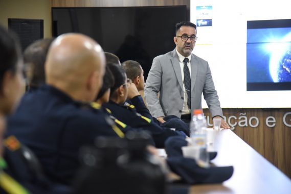 A imagem mostra uma palestra ou treinamento sendo ministrado em uma sala de situação e gerenciamento, voltada possivelmente a agentes de segurança pública. Descrição da cena: Um homem de terno cinza claro, óculos e gravata está em pé, gesticulando enquanto fala para um grupo de pessoas uniformizadas, indicando que é o palestrante ou facilitador da atividade. Os participantes estão sentados ao redor de uma mesa longa e retangular, usando uniformes escuros com insígnias e bandeiras do Brasil nos braços, sugerindo que são membros de alguma corporação — provavelmente policiais militares ou bombeiros. Sobre a mesa há copos descartáveis, garrafas de água e bonés do uniforme, indicando que é uma sessão relativamente longa e informal. Ao fundo, uma grande tela exibe uma apresentação com os termos “Mundo VUCA” e “Mundo BANI”, conceitos usados para descrever contextos modernos de instabilidade, incerteza e complexidade, bastante aplicados em ambientes de liderança e estratégia. Parte da frase visível na tela diz: “Como podemos definir ou entender nossa realidade atual?”, o que sugere que o tema da palestra está relacionado a cenários contemporâneos, tomada de decisão e adaptação em tempos de mudança. Ambiente: A sala possui acabamento moderno, com painéis de madeira e ar-condicionado. O letreiro ao fundo indica que é a “Sala de Situação e Gerenciamento”, reforçando o caráter tático e estratégico da reunião.