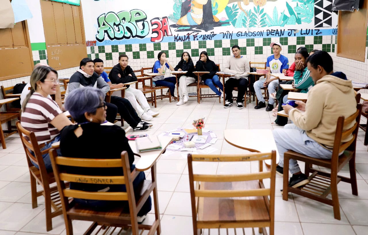 A imagem mostra uma roda de conversa em uma sala de aula. Os participantes, em sua maioria jovens, estão sentados em cadeiras organizadas em círculo, o que favorece o diálogo e a troca de ideias. No centro do círculo há uma pequena mesa improvisada com papéis, livros e uma flor decorativa, indicando um ambiente acolhedor e colaborativo. A sala tem paredes decoradas com azulejos verdes e brancos, além de um mural colorido com grafite que inclui palavras como "ACRE", "3A", "Kauã F. Jwayi", "WW Gladson" e "Dean Jr. Luiz". Todos parecem atentos e envolvidos, sugerindo que se trata de uma atividade pedagógica ou dinâmica de grupo com objetivo educativo ou reflexivo.
