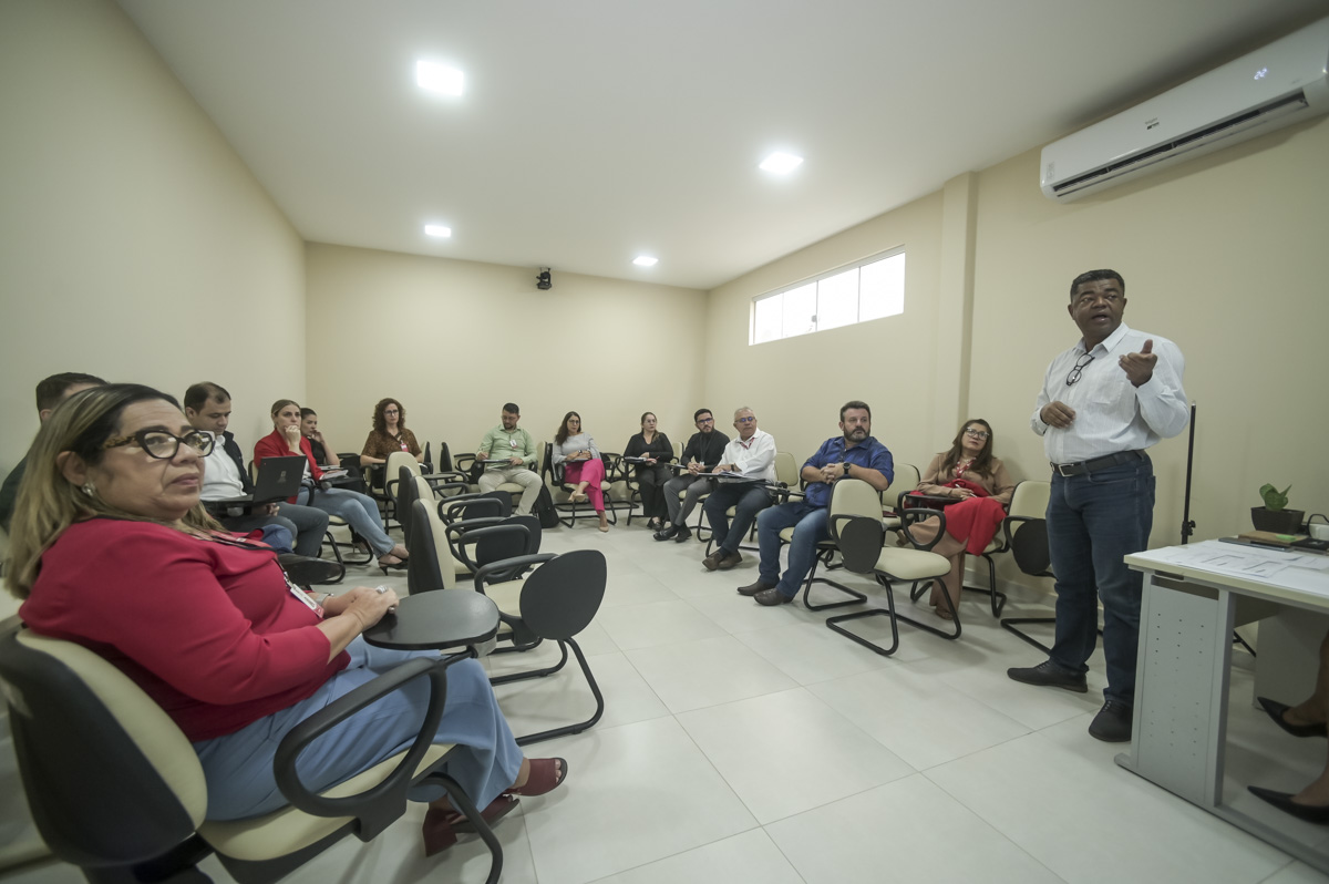 Uma imagem de grupo de pessoas em um ambiente de sala de aula ou reunião. A composição da imagem apresenta um grupo de pessoas sentadas em círculo, em cadeiras de plástico bege e pretas. O espaço é amplo, com paredes claras e piso claro. A imagem se concentra principalmente na disposição do grupo em círculo. Há uma pessoa em pé no centro do círculo, que parece estar apresentando ou conduzindo a reunião. O ambiente tem uma iluminação suave e uniforme, com uma aparência neutra. Os indivíduos demonstram um comportamento de escuta e participação, com posturas relaxadas mas atentas. Eles estão vestindo roupas casuais e de trabalho, com cores variadas. Há uma distribuição equilibrada de gênero e uma ampla variedade de idades. A imagem é fotográfica e tem uma aparência profissional e neutra. A técnica fotográfica foca na composição e nas pessoas em si, sem elementos adicionais ou recursos artísticos. O ambiente é uma sala de reunião ou aula. É uma sala interna bem iluminada, com paredes claras, piso de cerâmica e iluminação uniforme. A atmosfera é tranquila, profissional, sem efeitos de luz dramáticos ou foco em elementos externos. O clima parece neutro e focado em escuta e conversa.