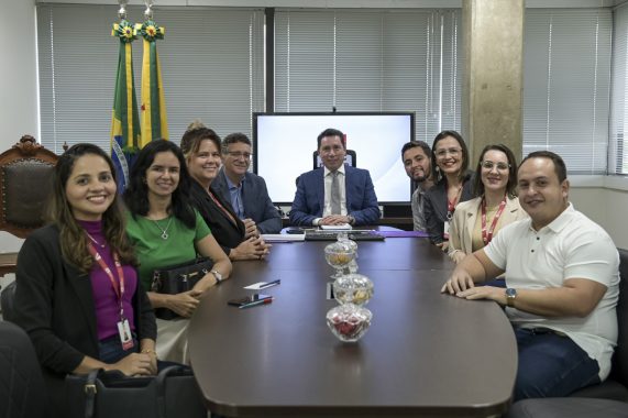 A imagem mostra um grupo de nove pessoas reunidas em torno de uma mesa de reunião em um ambiente corporativo. Todos estão sorrindo, indicando um momento de integração ou conclusão positiva de um encontro institucional. No centro da mesa há três potes de vidro com balas e petiscos. Ao fundo, há duas bandeiras do Brasil e do Piauí, reforçando que o local é uma instituição pública, provavelmente um órgão governamental ou do sistema judiciário. No centro, atrás da mesa, está um homem de terno azul e gravata clara, sentado em posição de destaque, possivelmente o anfitrião da reunião ou uma autoridade. As pessoas à mesa vestem trajes formais ou semi-formais, algumas com crachás, o que indica um ambiente profissional. Ao fundo, também se vê persianas fechadas cobrindo janelas amplas, além de uma tela ligada com um brasão institucional ao centro.