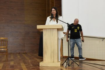 A imagem mostra uma mulher fazendo um discurso ou apresentação em um púlpito de madeira claro. Ela está vestida com um blazer branco e segura alguns papéis na mão esquerda, enquanto gesticula com a mão direita, transmitindo expressividade em sua fala. Há um copo com água sobre o púlpito, e um microfone direcionado para sua voz está posicionado em um suporte articulado. O fundo é dividido entre uma parede branca e uma parede com painéis de madeira escura, criando um ambiente formal e institucional, possivelmente relacionado a uma cerimônia, conferência ou evento público. A mulher aparenta estar concentrada e determinada, com uma postura firme e segura. O cenário reforça uma atmosfera de seriedade e profissionalismo.