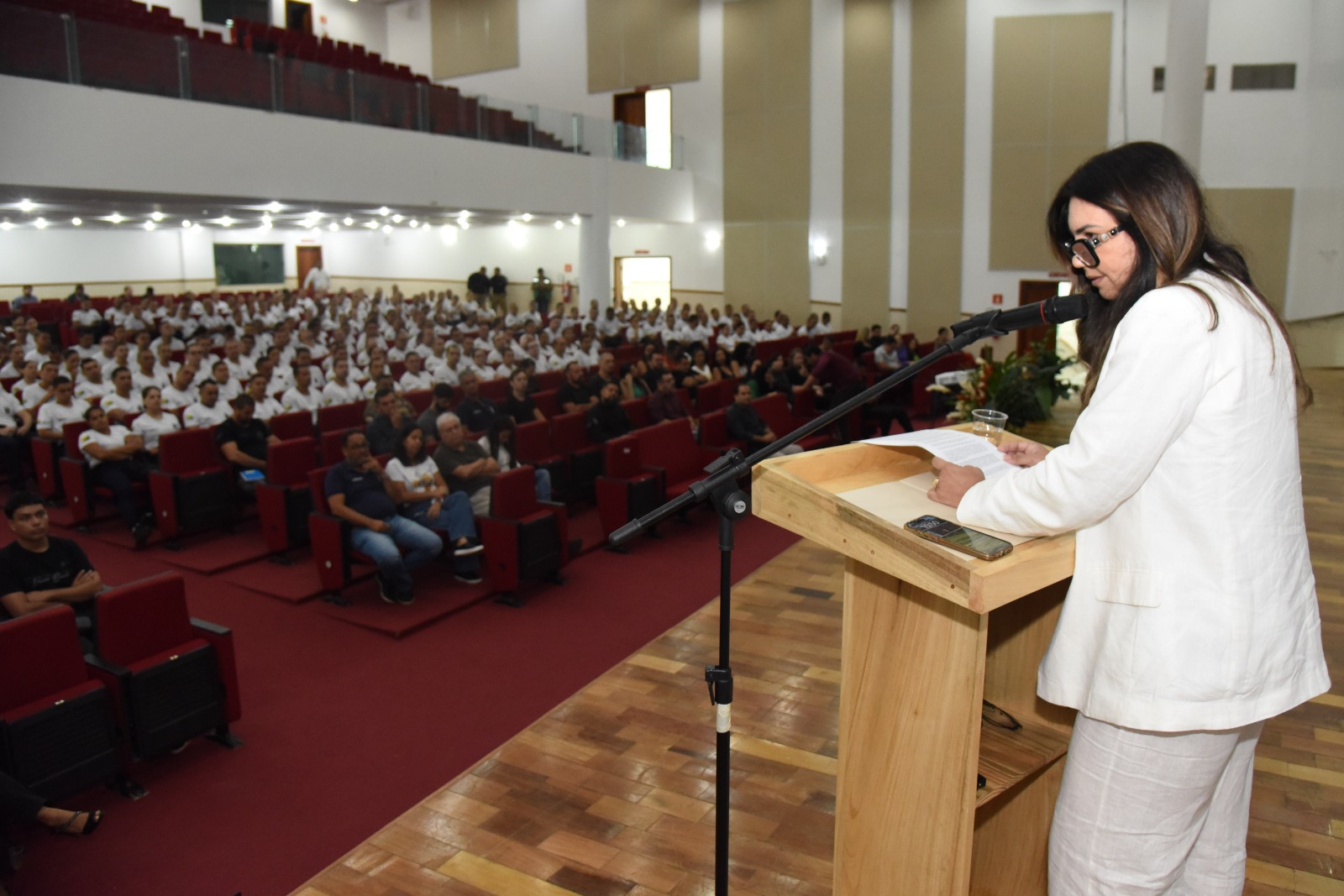 A imagem mostra uma mulher discursando em um auditório lotado. Ela está posicionada atrás de um púlpito de madeira, usando um terno branco e óculos escuros. A mulher segura uma folha de papel e fala ao microfone, demonstrando envolvimento com o público. O auditório é amplo, com assentos estofados vermelhos e ocupados por diversas pessoas, a maioria usando camisetas brancas — possivelmente participantes ou alunos de algum evento institucional ou formativo. Algumas pessoas também estão vestidas de forma mais casual, indicando a presença de convidados ou familiares. O piso é de madeira e há um carpete vermelho entre as fileiras de cadeiras. Ao fundo, nota-se uma galeria superior também com assentos. A cena transmite um ambiente formal, provavelmente relacionado a uma cerimônia, palestra ou evento educacional ou governamental. O celular sobre o púlpito e o copo com água reforçam a ideia de um discurso planejado e de certa duração.