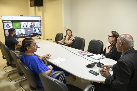 A imagem mostra uma reunião híbrida (presencial e virtual) ocorrendo em uma sala de reuniões simples, com paredes brancas e iluminação fluorescente. Seis pessoas estão fisicamente presentes, sentadas ao redor de uma mesa oval branca, e há uma tela grande exibindo uma videoconferência com vários participantes conectados remotamente. Detalhes observados: Presencialmente: À esquerda, um homem (de costas) está sentado operando ou observando a videoconferência. Uma mulher de tranças, vestindo azul, está sentada atenta, com celular e bloco de anotações à frente. Do outro lado da mesa, uma mulher de blusa estampada observa a tela com expressão pensativa, com a mão no queixo. Uma mulher com colar de pérolas e óculos (possivelmente a mesma da imagem anterior) observa atenta, com cadernos abertos à frente. Um homem grisalho de terno segura papéis e participa da conversa. Sobre a mesa, há microfones, cadernos, celular e outros materiais de apoio. Na tela: A videoconferência está sendo exibida em um monitor grande. Mostra vários participantes divididos em janelas, incluindo pessoas falando ou com câmeras ligadas. Ícones e botões da plataforma de videoconferência (provavelmente Google Meet) estão visíveis.