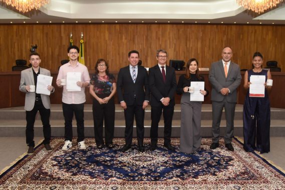 A imagem mostra um grupo de nove pessoas posando para uma foto em um ambiente formal, provavelmente um auditório ou plenário de um tribunal. O cenário tem um piso coberto por um tapete decorativo e um painel de madeira ao fundo com a presença de duas bandeiras oficiais (do Brasil e do estado), sugerindo uma solenidade institucional. Dos nove presentes: Cinco pessoas (três homens e duas mulheres) seguram canecas brancas e certificados/documentos, o que indica que estão sendo homenageados ou premiados. Os demais (quatro pessoas) estão posicionados no centro do grupo e aparentam ser autoridades ou organizadores do evento, vestindo trajes formais (ternos e roupas sociais). O clima da imagem é de celebração ou reconhecimento, como uma entrega de certificados, posse de estagiários, premiação ou conclusão de curso/formação no âmbito do Judiciário. A iluminação é clara e há dois lustres elegantes no teto, reforçando o ambiente solene.