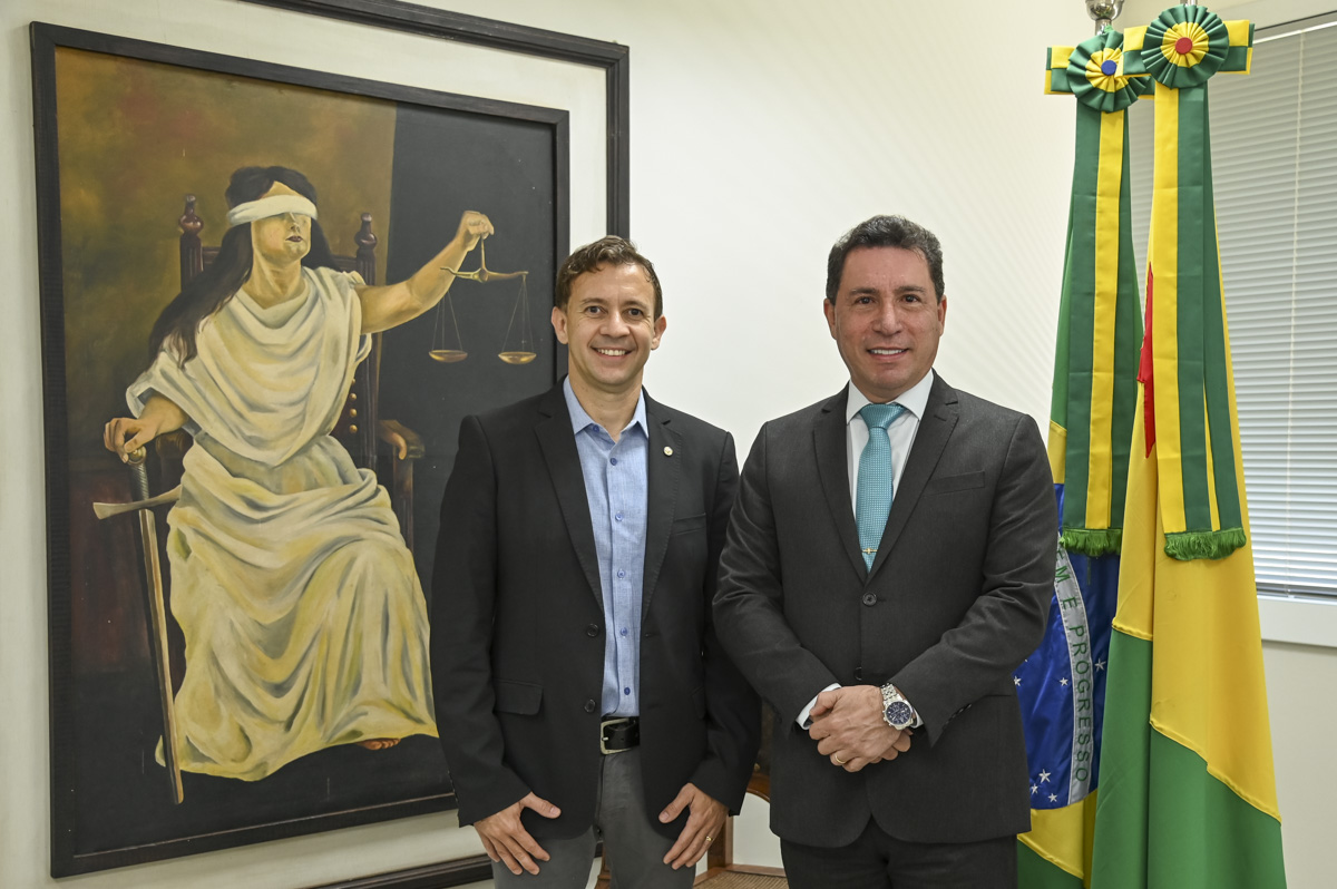 A imagem mostra dois homens em trajes sociais (terno e gravata), posando lado a lado para uma foto em um ambiente institucional, possivelmente um gabinete ou repartição pública. Ambos estão sorrindo levemente, transmitindo uma atmosfera cordial e formal. À esquerda da imagem, há uma grande pintura da deusa da justiça, representada com os olhos vendados, segurando uma balança em uma mão e uma espada na outra — símbolo clássico da imparcialidade e da justiça. À direita da imagem, estão visíveis duas bandeiras: a do Brasil e a do estado do Acre, ambas ornamentadas com laços cerimoniais, reforçando o contexto oficial da fotografia. O ambiente é claro e moderno, com persianas cobrindo as janelas ao fundo. A imagem sugere um momento de encontro institucional, possivelmente ligado ao Judiciário ou ao Poder Público.