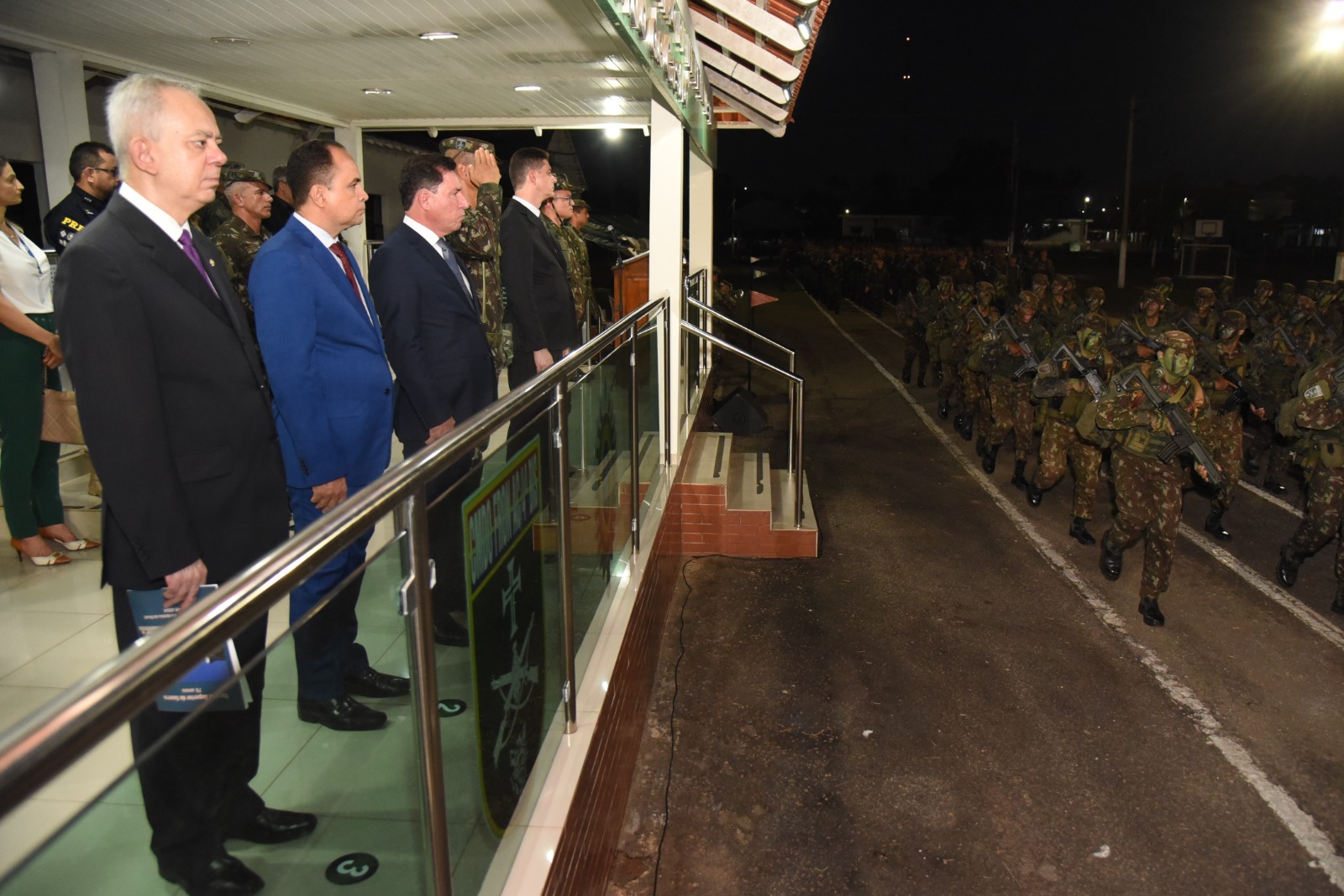 A imagem retrata uma cerimônia militar noturna em andamento, provavelmente em continuidade à solenidade mostrada na imagem anterior. No primeiro plano, autoridades civis e militares estão posicionadas em uma plataforma elevada e envidraçada, observando atentamente o desfile. Entre elas, destacam-se homens de terno — incluindo um em traje azul — e oficiais fardados, alguns dos quais prestam continência em sinal de respeito. À direita da imagem, um grande grupo de soldados do Exército Brasileiro marcha em formação pela pista asfaltada. Eles estão equipados com uniformes camuflados, capacetes e armamento, com rostos pintados de verde e preto, reforçando o caráter operacional da apresentação. A marcha ocorre com firmeza e sincronia, sob luzes artificiais que iluminam o local, indicando que o evento já ocorre durante a noite. Ao fundo, pode-se ver parte da estrutura do quartel e postes de iluminação, compondo o cenário típico de um ambiente militar. A solenidade parece integrar um evento oficial de alta relevância institucional, com a presença de autoridades e demonstração de tropa.