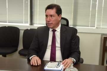 A imagem mostra um homem sentado à mesa em um ambiente corporativo ou institucional. Ele está usando um terno escuro, camisa branca e gravata vinho, transmitindo uma aparência formal e profissional. O homem olha para o lado, com uma expressão atenta, como se estivesse ouvindo ou participando de uma conversa ou reunião. Sobre a mesa à sua frente, há alguns itens pessoais e de trabalho, incluindo um caderno, um par de óculos, um smartphone com capa branca e uma caneta. Também há um objeto decorativo de vidro, semelhante a uma bombonière. Ao fundo, é possível ver cadeiras vazias com encosto acolchoado e persianas fechadas, sugerindo que a reunião ocorre em um espaço reservado e climatizado, típico de salas de reunião ou escritórios executivos. A cena passa uma sensação de seriedade e concentração, comum em ambientes institucionais.