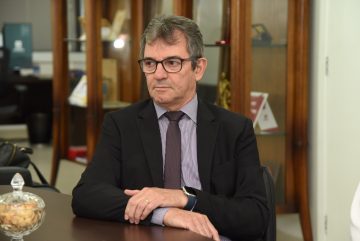 A imagem mostra um homem sentado à mesa durante o que parece ser uma reunião formal ou institucional. Ele está vestido com traje social: terno preto, camisa listrada e gravata escura. Usa óculos e um relógio inteligente no pulso esquerdo. Sua expressão é séria e atenta, indicando que está ouvindo ou refletindo sobre algo que está sendo discutido. Sobre a mesa à sua frente, há um pote de vidro com petiscos, o que é comum em ambientes de reunião. Ao fundo, há uma estante de madeira com portas de vidro, exibindo objetos como livros, troféus ou certificados — elementos típicos de um escritório ou sala de reuniões oficiais. O ambiente transmite um ar institucional, reforçado pela formalidade do traje e pelos itens decorativos e funcionais presentes na sala.