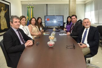 A imagem retrata uma reunião formal em uma sala corporativa ou institucional. O grupo, composto por sete pessoas — quatro mulheres e três homens — está posicionado ao redor de uma mesa de reuniões retangular, olhando para a câmera, o que indica que o registro foi feito de forma proposital, possivelmente para fins de divulgação institucional. Ao fundo, há duas bandeiras (do Brasil e do Acre), além de uma tela com uma apresentação digital exibindo o tema: “Julgamento ADPF 347 – Plano Pena Justa – Elaboração do Plano Estadual do Acre e Comitê de Políticas Penais”. Isso indica que a pauta da reunião está relacionada à política penitenciária e à implementação de um plano estadual voltado à justiça penal. As pessoas estão elegantemente vestidas, com trajes sociais, sugerindo que são profissionais de alto escalão ou autoridades públicas. Na mesa, há recipientes de vidro com balas e petiscos, além de cadernos, óculos, celulares e outros materiais típicos de reuniões executivas. O ambiente transmite seriedade e profissionalismo, reforçado pela presença de elementos simbólicos como as bandeiras, a apresentação institucional na TV e a pintura na parede com uma figura alegórica da Justiça.